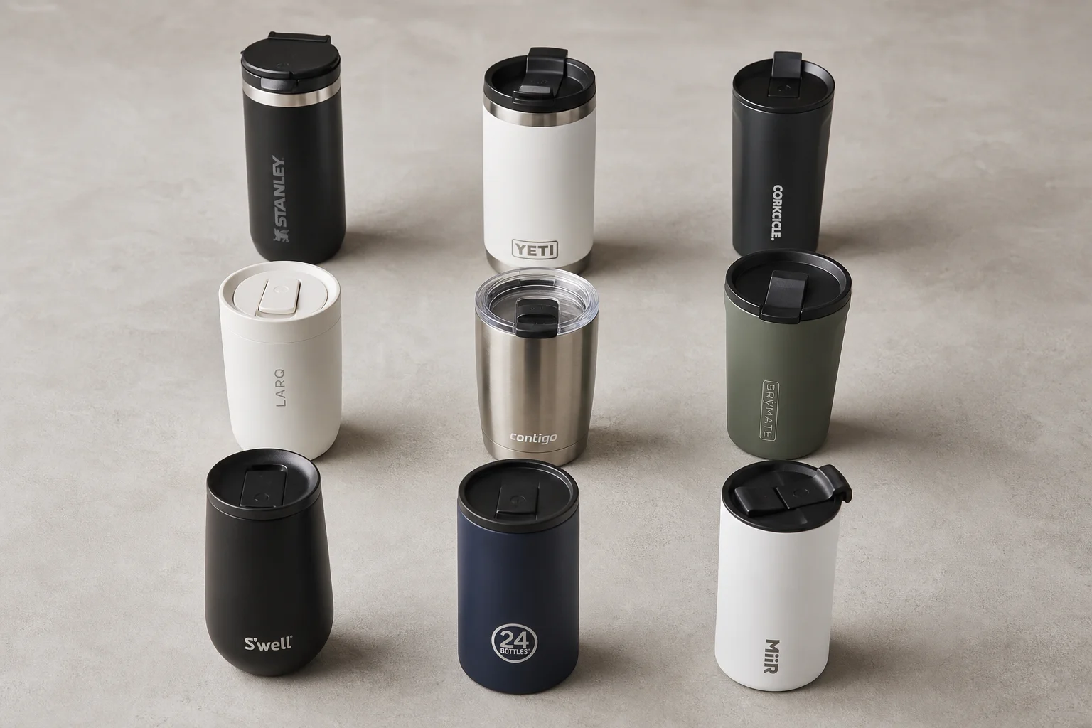 Nine insulated travel mugs arranged on concrete surface in various styles and colors