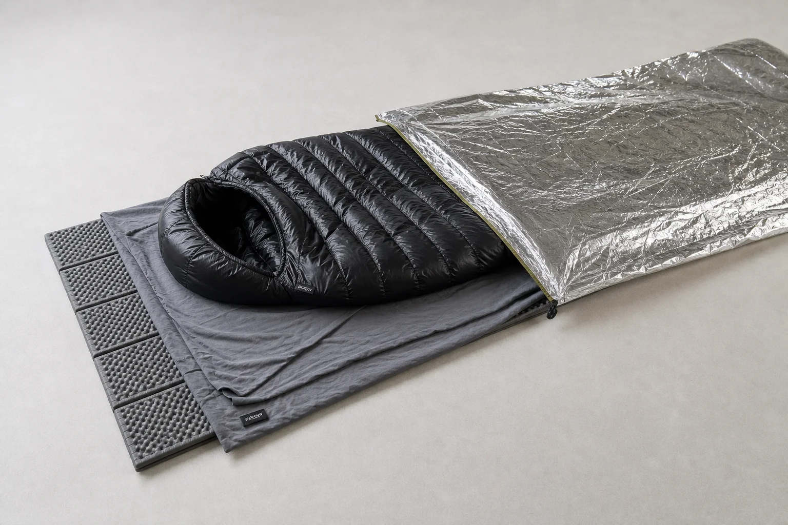 Layered sleep system components displayed flat showing mat, liner, bag, and bivvy