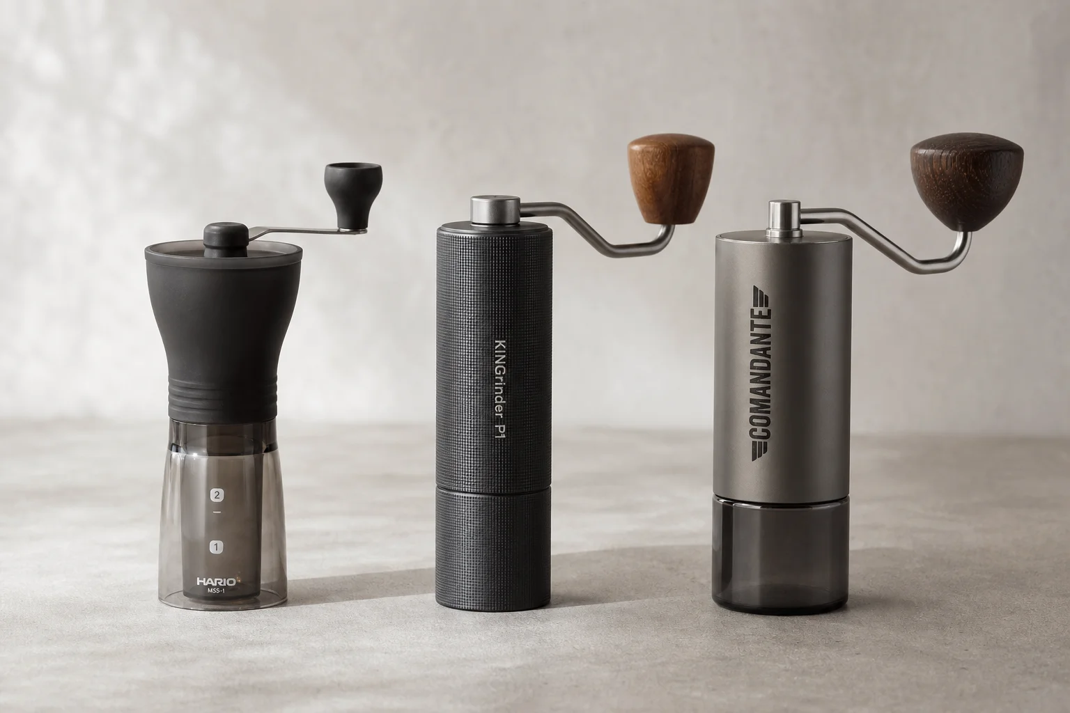 Three hand coffee grinders: Hario Slim, KINGrinder P1, and Comandante arranged on concrete
