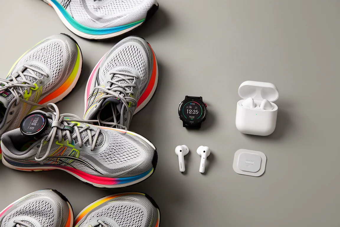 Flat-lay of racing shoes, GPS watches, earbuds and a sensor patch on a grey surface