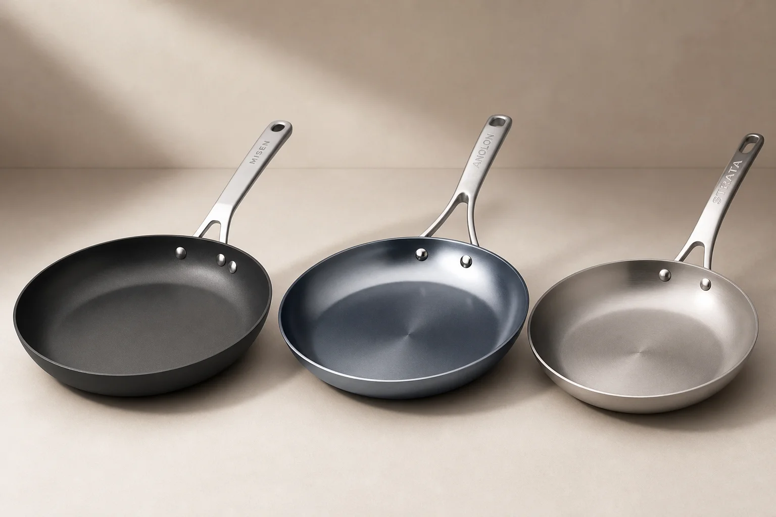 Tre nonstick-pannor i kolstål uppställda sida vid sida mot en neutral bakgrund