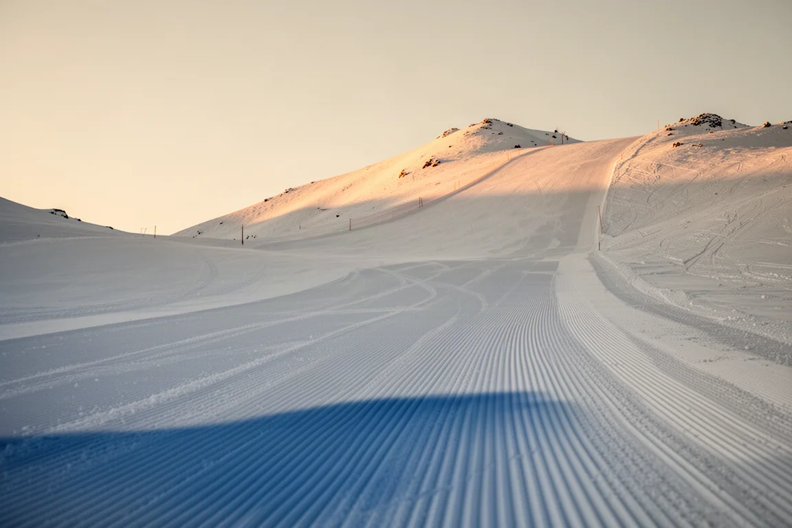 Piste de ski alpin fraîchement damée au lever du soleil, avec de fines stries de corduroy et des crêtes lointaines