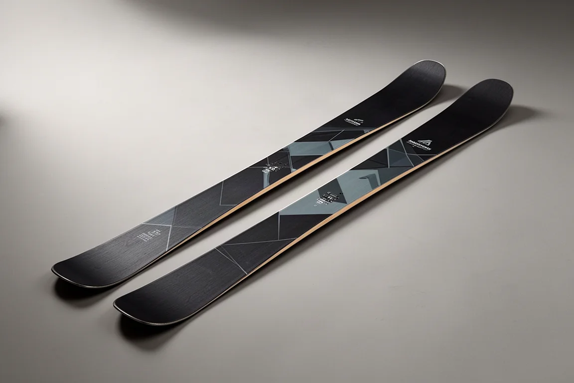 Pair of all-mountain skis with matte dark topsheets laid diagonally on a grey studio surface
