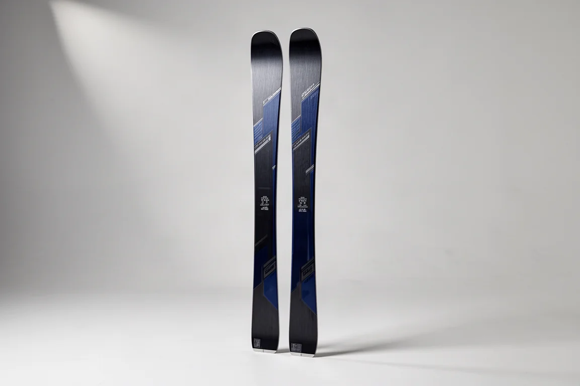 Par av smala frontside-alpinskidor med mörka grafit-belägg stående mot en ljusgrå bakgrund