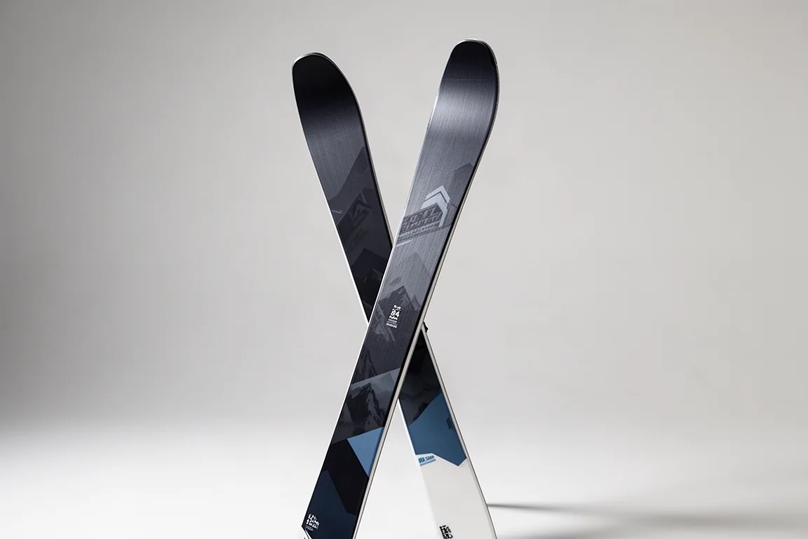 Pair of all-mountain skis with dark topsheets standing upright on a neutral grey backdrop